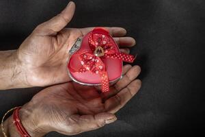 Female hands holding a heart shaped gift box with a red ribbon on a black background. Top view, close-up photo