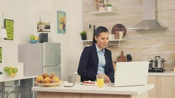 Jeune affaires femme dans le cuisine ayant une en bonne santé repas tandis que parlant sur une vidéo appel avec sa collègues de le bureau, en utilisant moderne La technologie et travail autour le l'horloge video