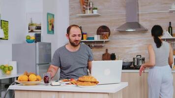 imprenditore Lavorando a partire dal casa mentre mangiare prima colazione nel cucina, indossare pigiama godendo arrostito pane con Burro. libero professionista Lavorando in linea attraverso Internet utilizzando moderno digitale tecnologia video