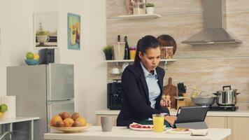 freelancer drinken koffie in de ochtend- Aan tafel top gedurende ontbijt gebruik makend van tablet computer. bedrijf vrouw lezing de laatste nieuws online voordat gaan naar werk, gebruik makend van modern technologie in de keuken terwijl aan het eten een gezond maaltijd video