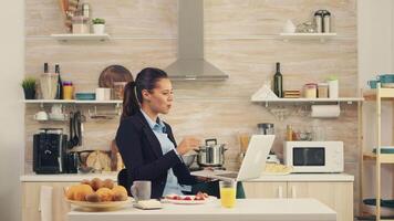 företag kvinna vinka på video ring upp under frukost. ung frilansare i de kök har en friska måltid medan talande på en video ring upp med henne kollegor från de kontor, använder sig av modern teknologi och arbetssätt runt om de klocka