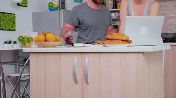 Happy couple surfing on internet using laptop during breakfast in kitchen. Married husband and wife in pajamas using internet web online modern technology, smiling and happy in the morning. Reading news browsing internet video