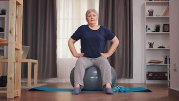 Senior woman doing fitness warm up on swiss ball in living room. Old person training home healthy retirement lifestyle stretching, home training and exercise video