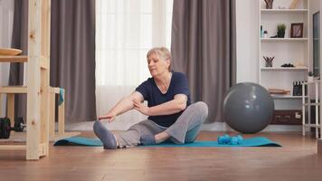 antiguo mujer extensión piernas sentado en yoga estera en vivo habitación. activo sano estilo de vida deportivo antiguo persona formación rutina de ejercicio hogar bienestar y interior hacer ejercicio video