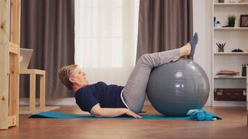 Senior woman training abs on swiss ball in living room. Old person healthy lifestyle pensioner living sport and wellness training indoors at home video