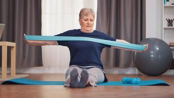 senior vrouw opleiding met weerstand band zittend Aan yoga mat. gepensioneerde oud vrouw uitrekken geschiktheid leven gezond levensstijl Bij pensioen, opleiding Bij huis oefening video