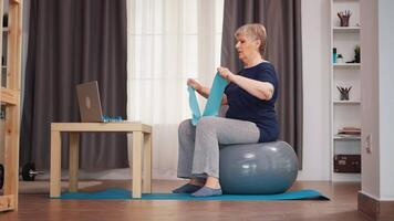 Sénior femme sur la stabilité Balle exercice avec la résistance bande en train de regarder en ligne leçon. en ligne formation apprentissage La technologie vieux femme levage formation en bonne santé mode de vie sport aptitude faire des exercices à Accueil avec poids haltère activité video