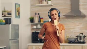 Cheerful woman singing on wooden spoon in the morning listening music on headphones. Energetic, positive, happy, funny and cute housewife dancing alone in the house. Entertainment and leiuse alone at home video