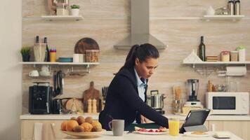 zakenvrouw drinken vers sap gedurende ontbijt voordat werk aan het kijken een video Aan tablet. bedrijf vrouw lezing de laatste nieuws online voordat gaan naar werk, gebruik makend van modern technologie in de keuken terwijl aan het eten een gezond maaltijd