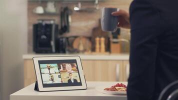 Business woman using smart device while eating breakfast in kitchen. Young freelancer at home talking on a video call with her colleagues from the office, using modern internet technology