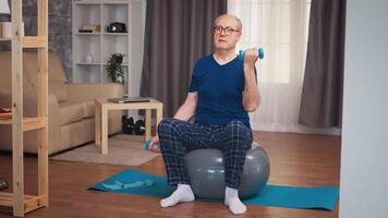 Old man doing biceps training with dumbbells on stability ball. Old person pensioner healthy training healthcare sport at home, exercising fitness activity at elderly age video