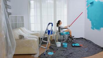 Paralyzed woman painting wall with roller brush sitting on wheelchair. Handicapped, disabled ill and immobilizes woman helping with apartment redecoration and home construction while renovating and improving. video