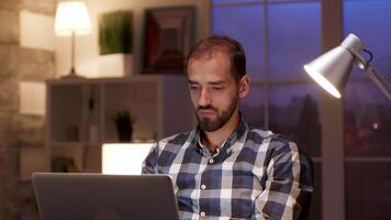 Bearded businessman stretching while working on laptop during night hours in home office. video