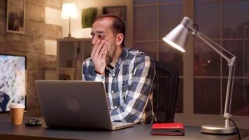 stanco manager sbadigli nel davanti di il suo il computer portatile mentre Lavorando nel casa ufficio durante notte ore. video