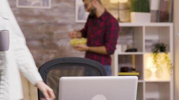 ragazza sorridente mentre Lavorando su il computer portatile nel vivente camera. fidanzato mangiare patatine fritte nel il sfondo. video