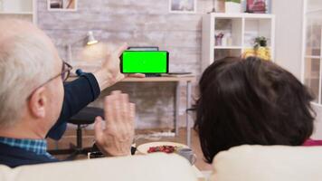 Back view of elderly age couple looking at phone with green screen while sitting on sofa in living room. video