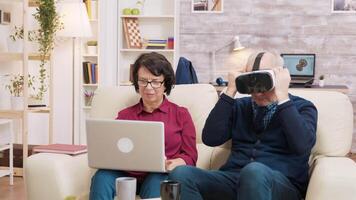 Senior man trying a VR headset in the living room while his wife uses a laptop next to him. Modern old couple using technology video