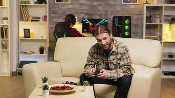 Excited man jumping in the air after his victory while playing video games using wireless controller. Girlfriend relaxing on computer in the background.