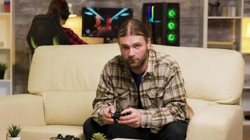 Focused bearded man sitting on couch playing video games using wireless controller. Girlfriend playing on computer in the background.