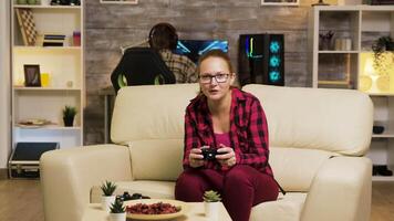 Woman sitting on couch in living room playing video games using wireless controllers. Boyfriend playing games on computer in the background.