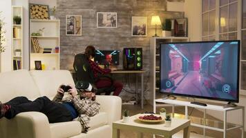 Man lying on sofa playing video games using vr headset with his girlfriend in the background playing on computer.