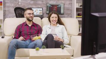 charmant jong paar zittend Aan de bankstel en aan het kijken TV terwijl genieten van hun chips. video