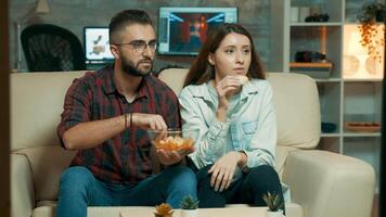 Concentrated caucasian young couple while watching tv and eating chips sitting on sofa. video