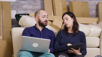 casal sentado em a chão do seus Novo apartamento e fazer conectados compras em computador portátil e tábua. video