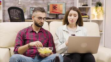 casal bebendo refrigerante e comendo salgadinhos enquanto navegando em computador portátil. casal relaxante em a sofá. video