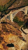 Sunlit rocks of the California coastline. Beautiful arched bridge connecting the mountains and highways on them. Aerial view. Vertical video