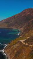 Brown mountains with highway at the coast of California. Craggy rocks descending into dark blue waters of Pacific Ocean. Top view. Vertical video