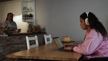 a woman in a pink jacket is sitting at a table with a laptop and a woman cooking at the background video