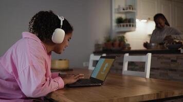 a woman in a pink jacket is sitting at a table with a laptop and a woman cooking at the background video