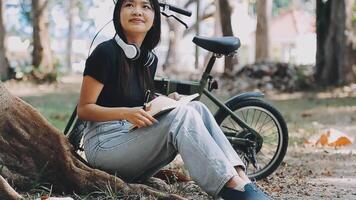 estilo de vida, educación, adolescencia. Adolescente estudiante en auriculares con teléfono inteligente bicicleta sentado en parque, hembra mira a teléfono pantalla video