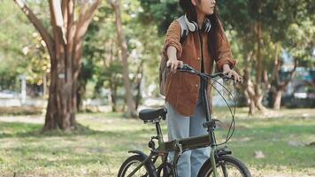 livsstil, utbildning, ungdom. Tonårs studerande i hörlurar med smartphone cykel Sammanträde i parkera, kvinna utseende på telefon skärm video