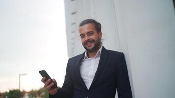 a man in a suit leaning on a wall is holding a cell phone video