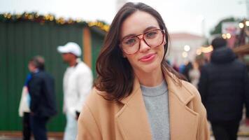 une femme portant des lunettes et une manteau dans une Noël marché est souriant à le caméra video