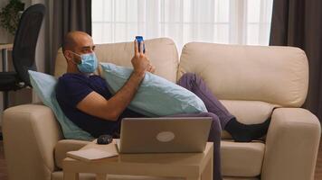 Man with protective mask laying on sofa running business from his phone during covid-19. video