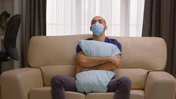 Sad man holding a pillow during coronavirus quarantine sitting on sofa wearing a mask. video