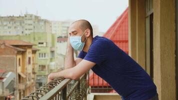 verdrietig Mens met beschikbaar masker op zoek ver weg van zijn balkon gedurende covid-19 pandemie. video