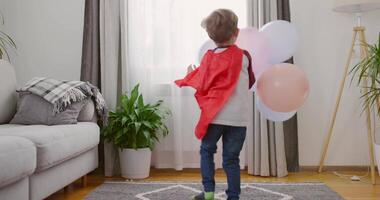 niño en superhéroe disfraz con rojo capa y máscara jugando con globos en hogar video