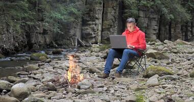 un hombre trabajos en un ordenador portátil cerca un corriente en naturaleza. concepto de trabajo independiente, digital nómada o remoto oficina. 4k video