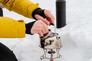 Hand holds kettle, open kettle lid, brew coffee, cook food in the woods, tourist utensils, metal kettle stands on gas burner, mountaineering equipment photo