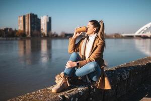 Beautiful woman in warm clothing enjoys drinking coffee and resting by the river on a sunny winter day.Toned image, photo