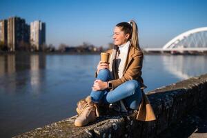 Beautiful woman in warm clothing enjoys drinking coffee and resting by the river on a sunny winter day. photo