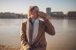 Beautiful woman in warm clothing enjoys drinking coffee and resting by the river on a sunny winter day.Toned image, photo