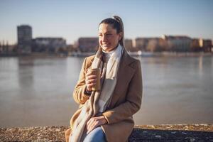 Beautiful woman in warm clothing enjoys drinking coffee and resting by the river on a sunny winter day. Toned image. photo