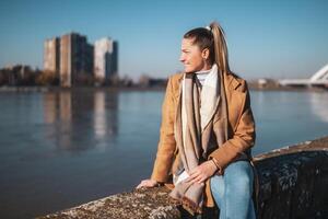 Beautiful woman in warm clothing enjoys resting by the river on a sunny winter day photo