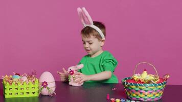 adorable peu enfant en jouant avec une farci lapin et une rose Oeuf à tableau, ayant amusement décorer de fête Pâques ornements dans studio. de bonne humeur content garçon profiter vacances les préparatifs. caméra un. video