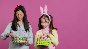 froh zuversichtlich Kind und Mutter zeigen Ostern Körbe auf Kamera, dekorieren festlich Vereinbarungen zum Frühling Urlaub. glücklich Schülerin mit Hase Ohren posieren mit ihr Mama im Studio. Kamera a. video